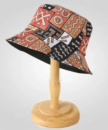 Reversible bucket hat with vibrant African-inspired geometric and abstract print in black, orange, brown, and white. Shown on a wooden stand.