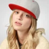 A woman with blonde hair wears a gray flat-bill hat with a red brim.